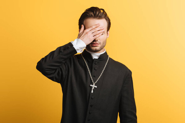 young catholic priest covering eyes with hand isolated on yellow