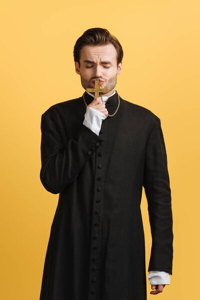 young faithful catholic priest kissing cross with closed eyes isolated on yellow