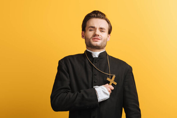 young catholic priest with exhausted face expression holding cross near heart isolated on yellow