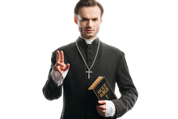 confident, strict catholic priest holding holy bible and showing blessing gesture isolated on white