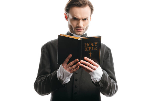 frowning catholic priest looking at camera while holding holy bible isolated on white
