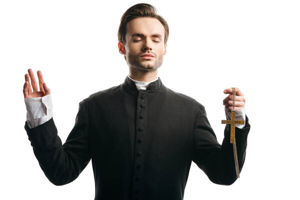 concentrated catholic priest praying with raised hand while holding golden cross isolated on white