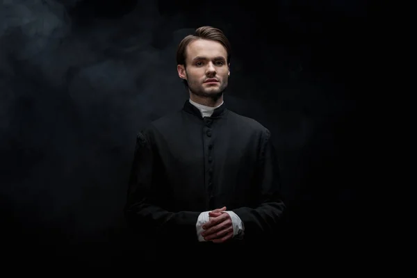 Young Serious Catholic Priest Standing Folded Hands Black Background Smoke — Stockfoto