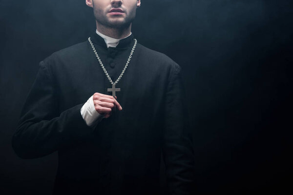 cropped view of catholic priest touching silver cross on his necklace on black background with smoke