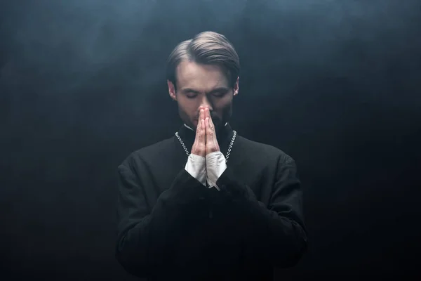 Young Concentrated Catholic Priest Praying Closed Eyes Black Background Smoke — Stock Photo, Image