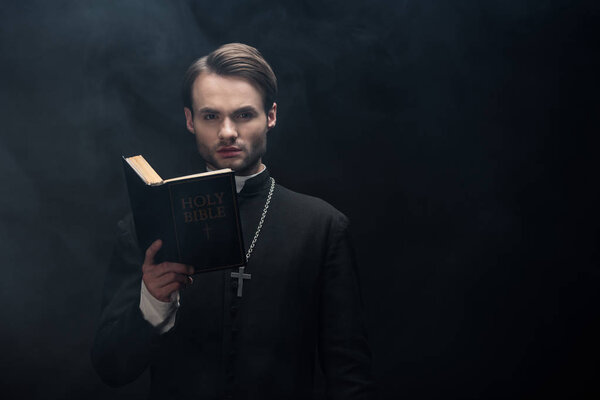 young serious catholic priest looking at camera while reading bible on black background with smoke