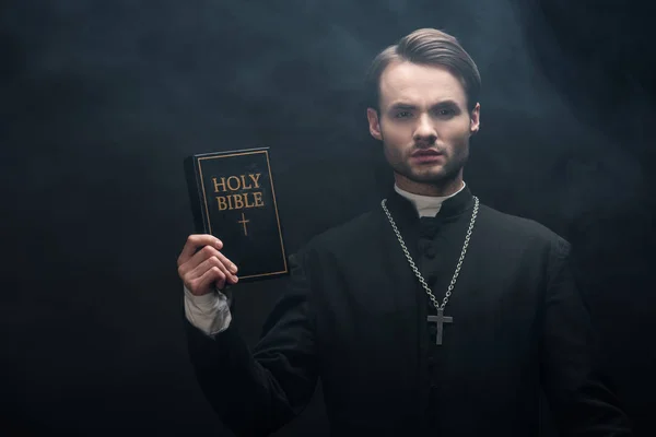 Młody Poważny Katolicki Ksiądz Pokazując Świętą Biblię Przed Kamerą Czarnym — Zdjęcie stockowe