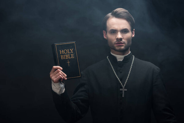 young serious catholic priest showing holy bible at camera on black background with smoke