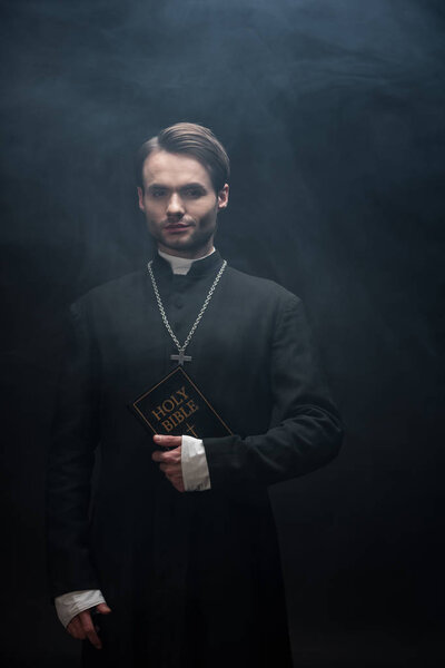 confident catholic priest holding holy bible and looking at camera on black background with smoke