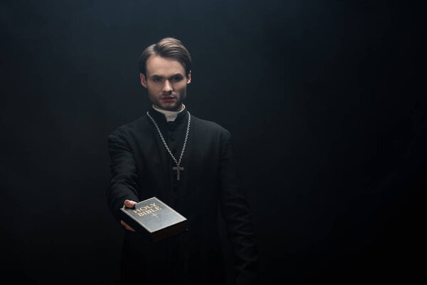 молодой серьезный католический священник держит священную Библию в протянутой руке изолированы на черный
