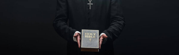cropped view of catholic priest holding holy bible isolated on black, panoramic shot
