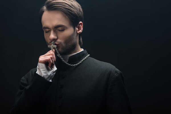 молодой вдумчивый католический священник целует серебряный крест с закрытыми глазами, изолированными на черном
