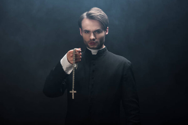 confident catholic priest holding necklace with cross while looking at camera on black background with smoke