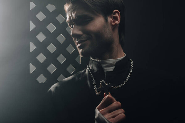 young worried catholic priest touching cross on his necklace in dark near confessional grille