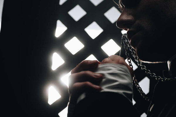 обрезанный вид католического священника целующего крест на своем ожерелье рядом с исповедальней решеткой в темноте с лучами света
