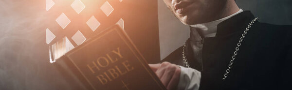 cropped view of catholic priest reading bible in dark near confessional grille with rays of light, panoramic shot