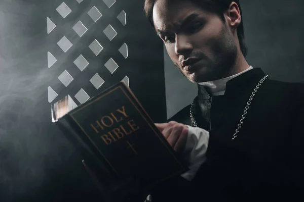 年轻体贴的天主教牧师 在黑暗中 带着光芒 靠近忏悔会的格栅阅读圣经 — 图库照片