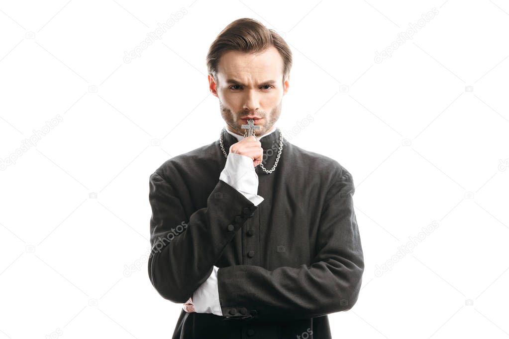 confident, serious catholic priest kissing holy cross while looking at camera isolated on white