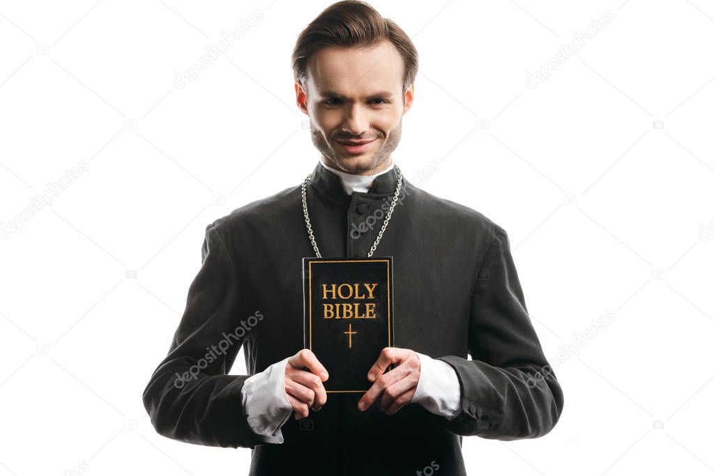 young, smiling catholic priest showing holy bible isolated on white