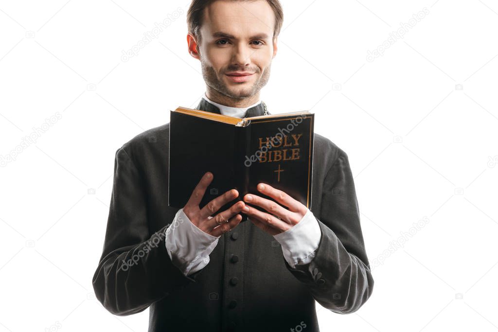 young, smiling catholic priest holding holy bible while looking at camera isolated on white