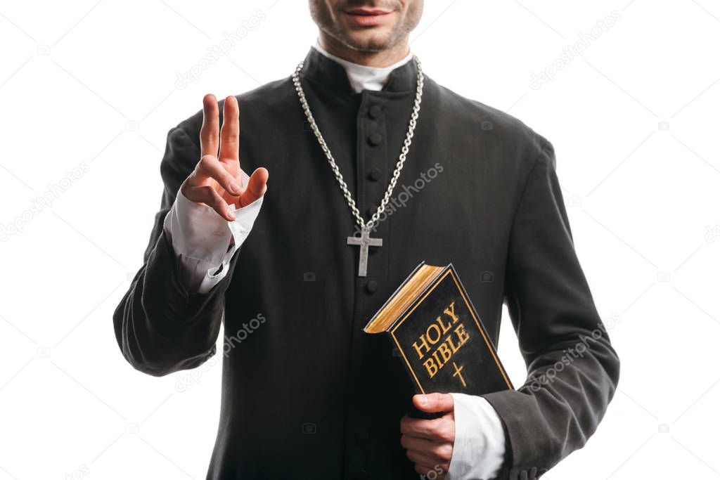 cropped view of young catholic priest holding holy bible and showing blessing gesture isolated on white