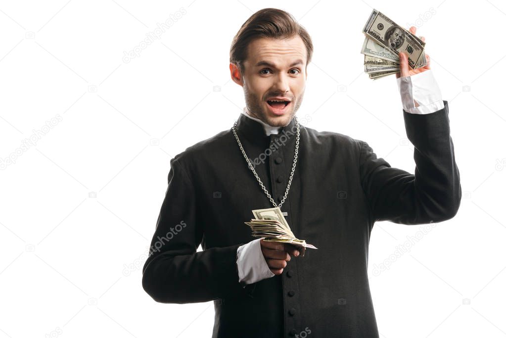 excited catholic priest looking at camera while showing dollar banknotes isolated on white