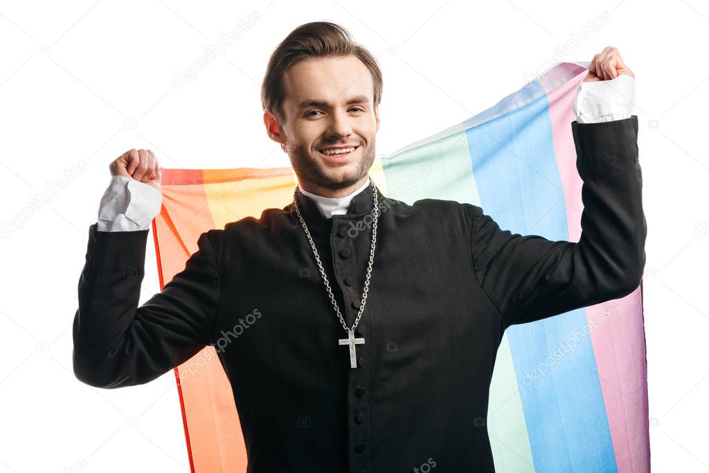 smiling catholic priest looking at camera while holding lgbt flag isolated on white