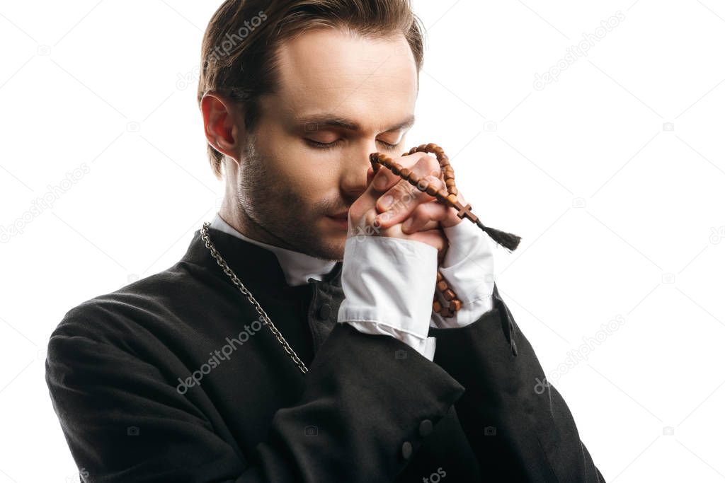 young, concentrated catholic priest praying with closed eyes while holding wooden rosary beads near face isolated on white