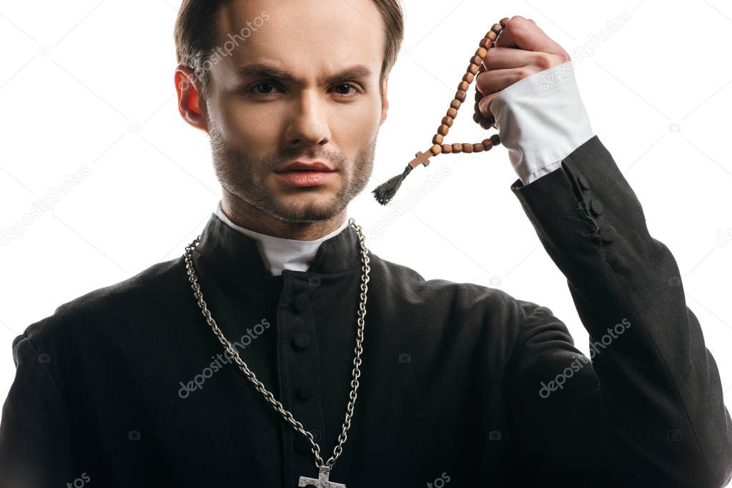 young, serious catholic priest holding wooden rosary beads while looking at camera isolated on white