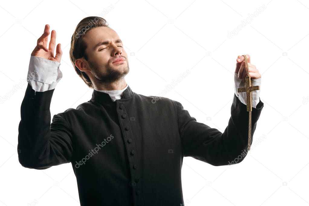 young catholic priest praying with closed eyes and raised hands while holding golden cross isolated on white