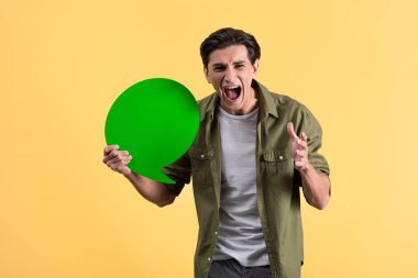 aggressive man shouting and holding green speech bubble, isolated on yellow