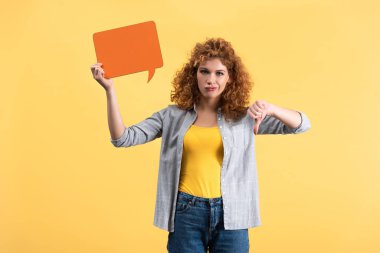 skeptical woman showing thumb down and holding empty speech bubble, isolated on yellow 