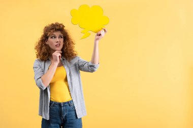 beautiful skeptical woman looking empty speech bubble in shape of cloud, isolated on yellow 