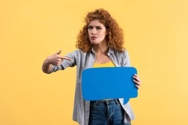 serious woman pointing at empty speech bubble, isolated on yellow 