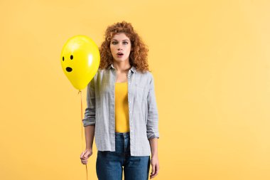 surprised redhead girl holding balloon with shocked face, isolated on yellow