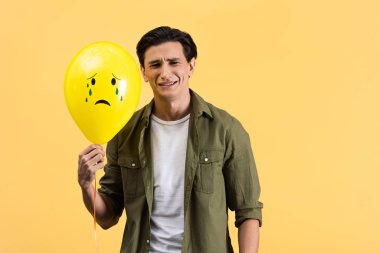 sad young man holding crying balloon with tears, isolated on yellow