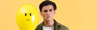 panoramic shot of skeptical man holding angry balloon, isolated on yellow