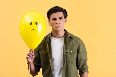 skeptical young man holding angry balloon, isolated on yellow