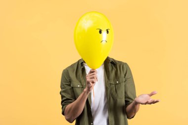 young man with shrug gesture holding angry balloon in front of face, isolated on yellow