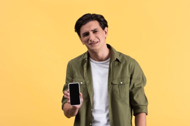 Stresli genç adam boş ekranlı akıllı telefon gösteriyor, sarı ekranda izole edilmiş.