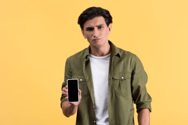 ciddi bir genç adam beyaz ekranlı akıllı telefon gösteriyor, sarı ekranda izole edilmiş.