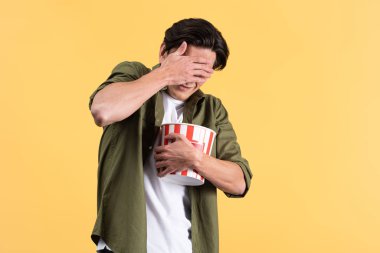 scared young man closing eyes while watching horror movie with bucket of popcorn, isolated on yellow