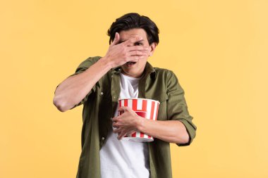frightened young man watching horror movie, closing eyes and holding bucket of popcorn, isolated on yellow