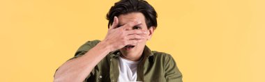 panoramic shot of scared young man watching horror movie and closing eyes, isolated on yellow