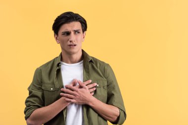 portrait of worried young man holding hands on heart, isolated on yellow