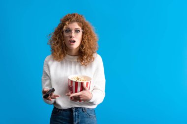 shocked girl watching tv with remote control and bucket of popcorn, isolated on blue