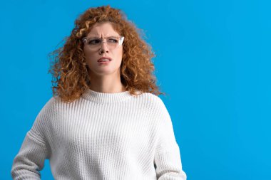 redhead skeptical girl in eyeglasses, isolated on blue