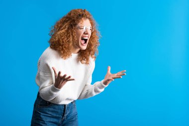 angry shouting redhead girl in eyeglasses, isolated on blue