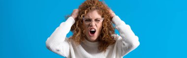 panoramic shot of aggressive screaming redhead girl in eyeglasses, isolated on blue
