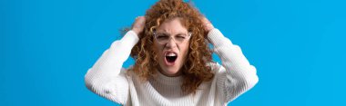 panoramic shot of aggressive screaming redhead girl in eyeglasses, isolated on blue clipart